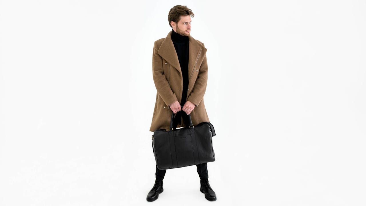 Fashionable man in coat holding a duffel bag in a minimalist studio setting.